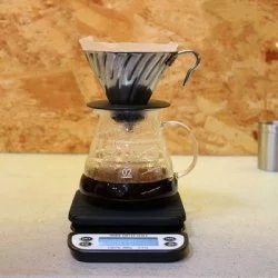 Baristická váha od Rhinowares v procesu přípravy kávy přes V60 se skleněnou konvičkou s dřevěným pozadí