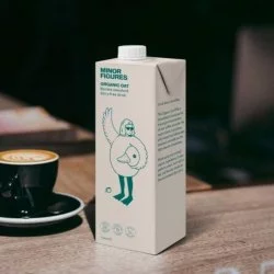Organické ovesné mléko zančky Minor Figures Organic Oat na dřevěném stole a vedle ležící cappuccino v černém porcelánovém šálku