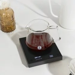 Černá váha s časovačem ležící na bílém stole společně se skleněnou konvicí s připravenou kávou