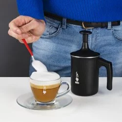 Černý napěňovač mléka zančky Bialetti Tuttocrema 330ml a v pozadí barista který lžící přidává našlehané mléko do Cappuccina