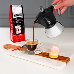 ukázka servírování kávy z hliníkové moka konvičky italské značky Bialetti do průhledného šálku na dřevěné prkýnko s pytlíkem kávy a malými makronkami