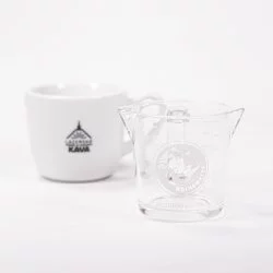 Skleněná odměrka na double espresso pro baristy značky Rhinowares Double Spout Shot Glass vedle šálku Lázeňské kávy