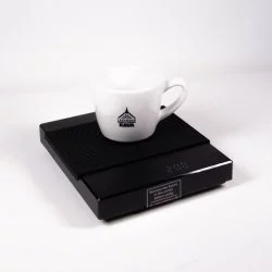 Černá baristická digitální váha Black Mirror s černou gumovou podložkou a na ní položený šálek lázeňské kávy