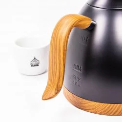 Detailní záběr na úchyt konvice značky Brewista v matně černém provedení s dřevěnými prvky pyšnící se pohodlnou manipulací, v pozadí šálek s logem Lázeňské kávy