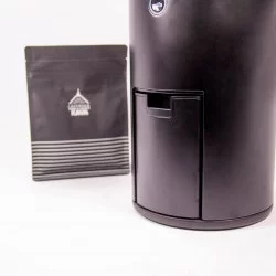 Detail na elektrický mlýnek na kávu zsnčky Wilfa Uniform WSFBS-100B společně s čerrným balíčkem s motivy Lázeňské kávy