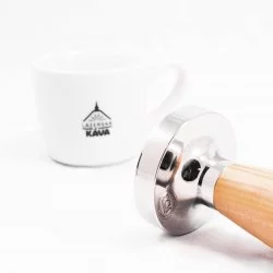 Detail tamperu s rukojetí z olivového dřeva s lázeňskou kávou v pozadí.