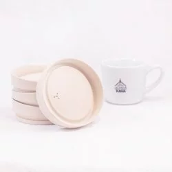 3 ekologická béžová víčka Huskee naskládaná na sobě, jedno o ně opřené a v pozadí šálek lázeňské kávy