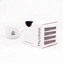 Černé ekologické víčko Huskee v originálním balení na bílém pozadí s šálkem lázeňské kávy