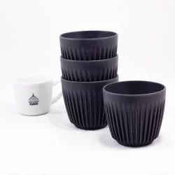 Ekologické thermo hrnky v černé barvě značky Huskee Charcoal na bílém stole s porcelánovým šálkem značky Lázeňská káva na bílém pozadíá