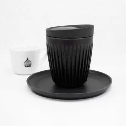 Černý termo hrnenk s podšálkem a víčkem značky Husky Charcoal o objemu 240ml na bílém stole a v pozadí šálek s motivy Lázeňské kávy