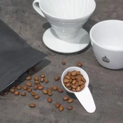Rozsypaná kávová zrnana kuchyňské desce s bílým dripperem a šálkem lázeňské kávy