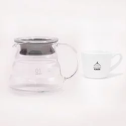 Skleněná konvička pro přípravu kávy V60 značky Hario se speciální pryžovou vložkou a v pozadí bílý šálek s logem Lázeňské kávy
