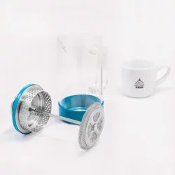 Skleněný French press v tyrkysové barvě snadnon rozložitelný a v pozadí porcelánový šálek s logem Lázeňská káva
