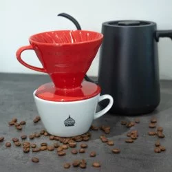 Černá elektrická konvice s šálkem lázeňské kávy na kterém leží červený dripper. Vše na šedé lince s rozsypanými kávovými zrny