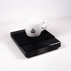 Černá baristická digitální váha Black Mirror s černou gumovou podložkou a na ní položený šálek lázeňské kávy Černá baristická digitální váha Black Mirror s černou gumovou podložkou a na ní položený šálek lázeňské kávy