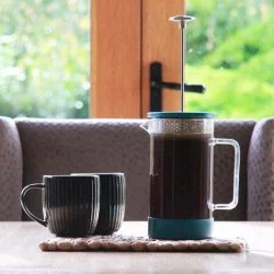 Proces přípravy kávy v modrém French pressu na bílém stole se dvěma černými šálky na kávu s výhledem do přírody