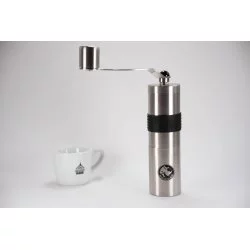 Ruční nerezový mlýnek na kávu na bílém stole s bílým hrnek značky Lázeňská káva