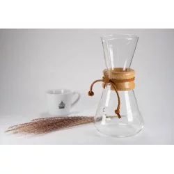 Skleněný Chemex s protáhlou hlavou dřevenou rukojetí a koženám provázkem bílý šálek na kávu rozprášená káva na bílem stole