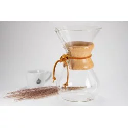 Skleněný Chemex s dřevěnou rukojetí s koženým provázkem bílý hrníček na kávu a rozprášená káva na bílém stole