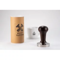 Heavy Temper o průměru 58 mm s černou rukojetí na bílém stole s šálkem Lázeňské kávy a originálním obalem pro temper