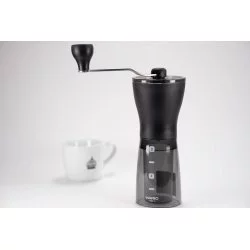 Černý ruční mlýnek na kávu na bílém stole a bílý hrnek na kávu značky Lázeňské kávy v pozadí