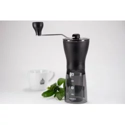 Černý ruční mlýnek na kávu na bílém stole s rostlinou a bílým šálkem na kávu zančky Lázeňská kává v pozadí