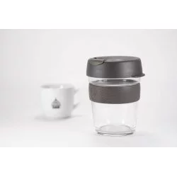 Skleněný termohrnek o objemu 340 ml s šedým víčkem a šedým gumovým držákem na bílém pozadí s šálkem lázeňské kávy